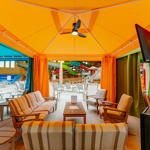 The image shows a colorful cabana with seating, a TV, and curtains, located near a water park with slides in the background.