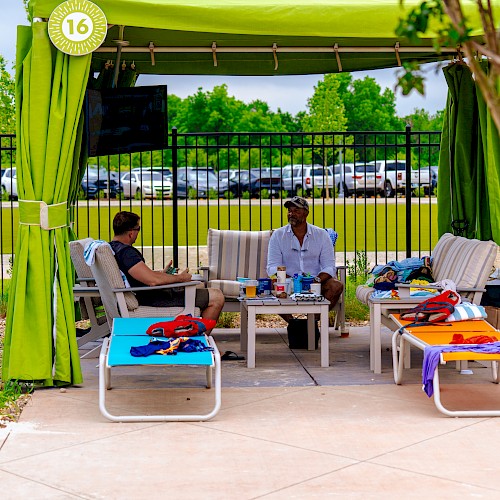 Two people sit in a green tented cabana with lounge chairs and a table. Bright towels are scattered around in a sunny outdoor setting.