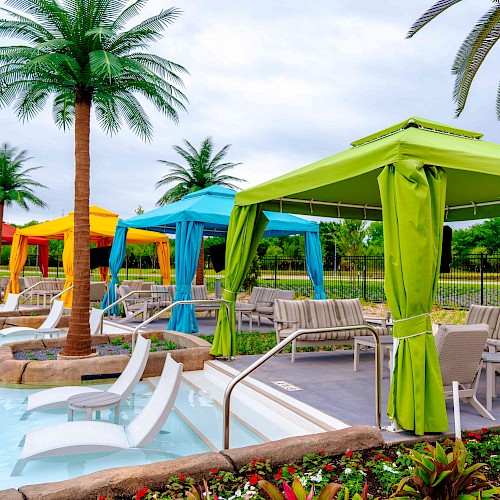 A vibrant pool scene with colorful cabanas, lounge chairs, and artificial palm trees, set against a landscaped backdrop.