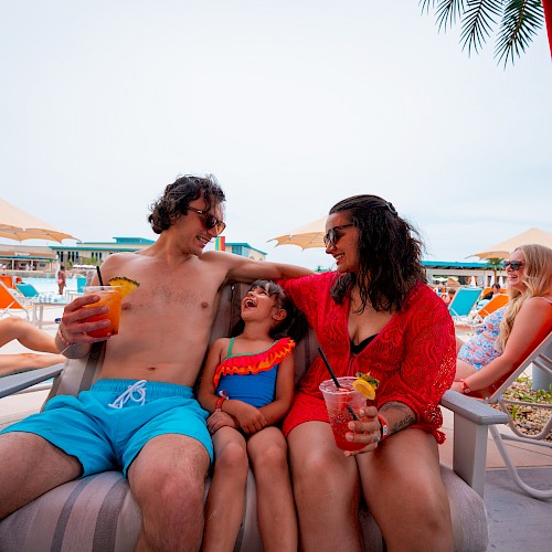 A family relaxes by the pool, enjoying drinks and sunshine, while people lounge in the background with umbrellas.