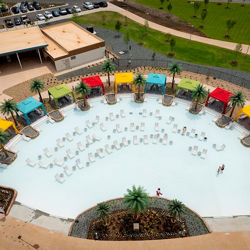 An aerial view of a circular pool surrounded by colorful cabanas and palm trees, with chairs arranged in the shallow water below.